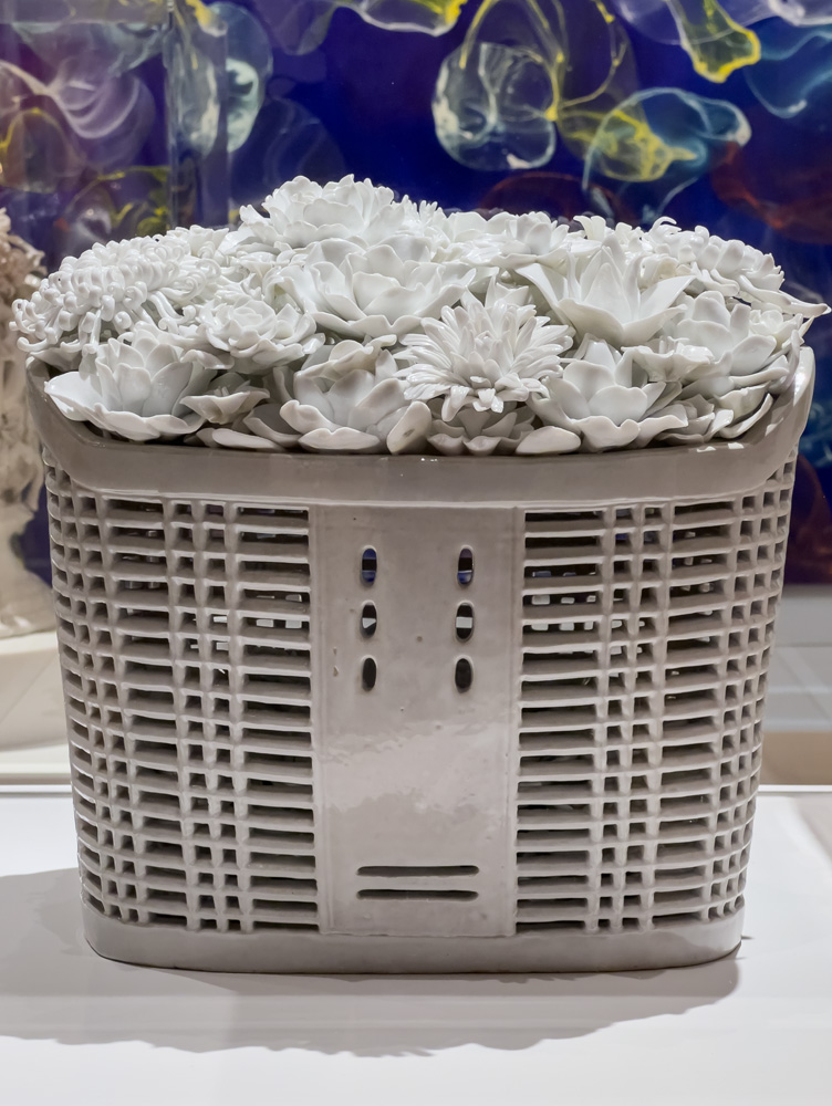 Ai Weiwei, Bicycle Basket with Flowers, 2014, Porcelain 11 x 14 1/2 by 13 1/2 inches (28 x 37 x 34 cm) with Jiří Georg Dokoupil, Untitled #4, 2015 in the background. Bicycle Basket with Flowers is Weiwei's porcelain reference to his 600-day brilliant silent social media protest "With Flowers." It was his artistic response to his travel ban by the Chinese government. This colorless bouquet of flowers stands in memoriam of the artist's loss of a right to life, freedom, and humanity. Ai states, "Expressing oneself is a part of being human. To be deprived of a voice is to be told you are not a participant in a society; ultimately, it is a denial of humanity."